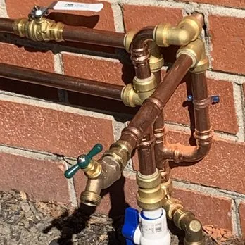 A detailed image of copper pipes and valves connected against a brick wall, showcasing plumbing installation by Puronics, LLC in Livermore.